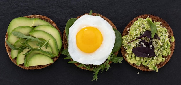 Avocado sandwich on dark rye bread with spinach and fried egg on a dark background, top view.