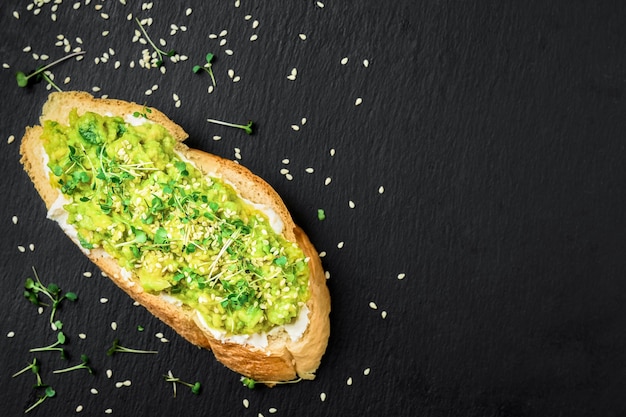 Avocado sandwich with olive oil, microgreens and sesame seeds on baguette toast with cream cheese on a black table. Top view with copy space