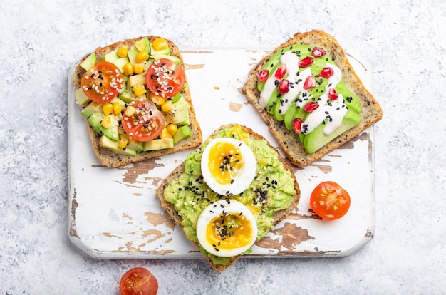 Avocado toast met ei, tomaten, kruiden over witte houten rustieke snijplank op stenen achtergrond. Gezonde ontbijtavocado sandwiches maken met verschillende toppings, bovenaanzicht, close-up