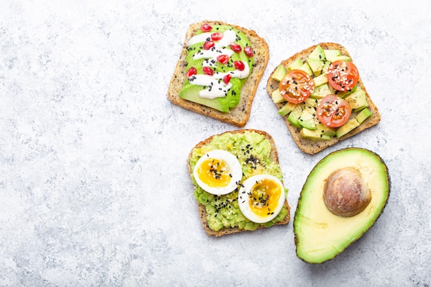 Avocado toasts with egg, tomatoes, seasonings and a half of whole avocado over white stone background, space for text. Healthy breakfast avocado sandwiches with different toppings, top view, close-up