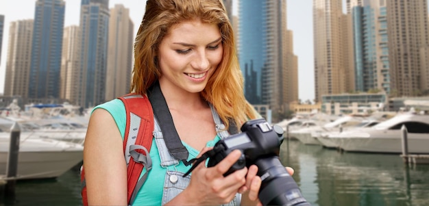 avontuur, reizen, toerisme, wandeling en mensenconcept - gelukkige jonge vrouw met rugzak en camera die over de stad en de haven van Dubai fotografeert met botenachtergrond