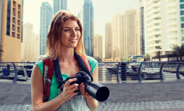 avontuur, reizen, toerisme, wandeling en mensenconcept - gelukkige jonge vrouw met rugzak en camera die over de stad en de haven van Dubai fotografeert met botenachtergrond