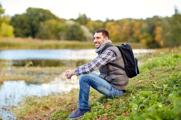 avontuur, reizen, toerisme, wandeling en mensenconcept - glimlachende man met rugzak die op de oever van de rivier rust