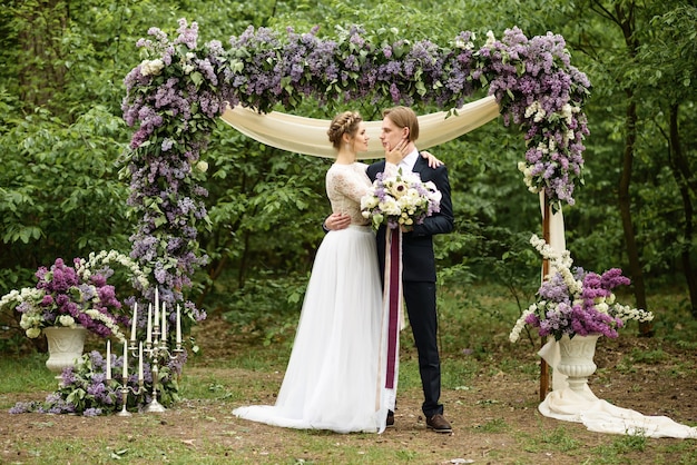 away wedding ceremony in the woods. the bride and groom stand near the lilac flower arch
