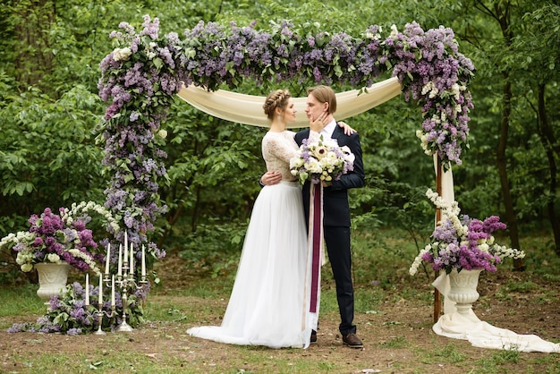 Away wedding ceremony in the woods. The bride and groom stand near the lilac flower arch