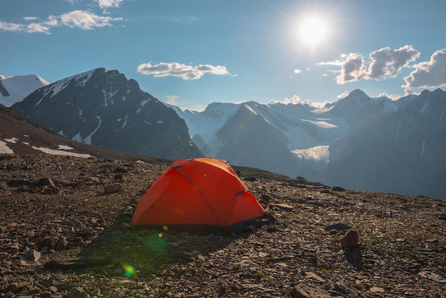 Awesome alpine landscape with orange tent with view to high snow mountains and large glacier Bright sun in cloudy sky above snowy mountain range Tent with top view to glacier tongue shining in sun