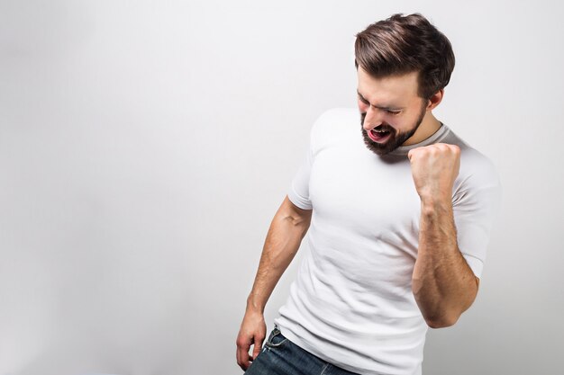 Awesome and beautiful guy is standing at the right side of a frame and showing that he is happy. He has made a fist out of his hand and looking down to the floor. Isolated on white wall