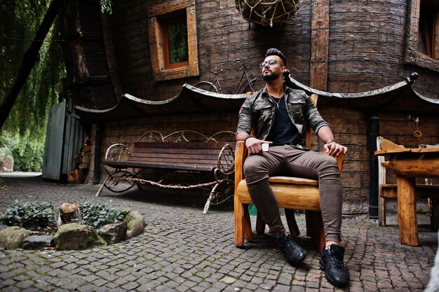 Awesome beautiful tall ararbian beard macho man in glasses and military jacket posed outdoor sitting on chair against wooden house