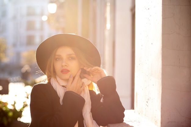 Awesome blonde woman wearing coat and hat posing at the old street. Space for text