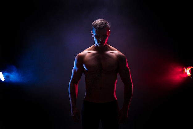 Awesome bodybuilder silhouette. Handsome power athletic man bodybuilder. Fitness muscular body on dark colour smoke background. Perfect male. tattoo, posing. 