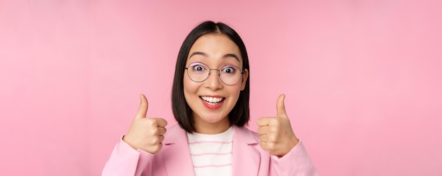 Удивительные поздравления Лицо взволнованной азиатской деловой женщины в очках, улыбающейся, довольной, показывая большие пальцы в знак одобрения, стоящей на розовом фоне