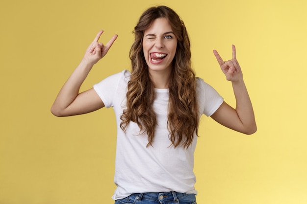 Awesome party having fun. Daring attractive carefree girl enjoy excellent event show rock-n-roll heavy metal gesture mimicking funny expressions wink stick tongue stand yellow background amused.