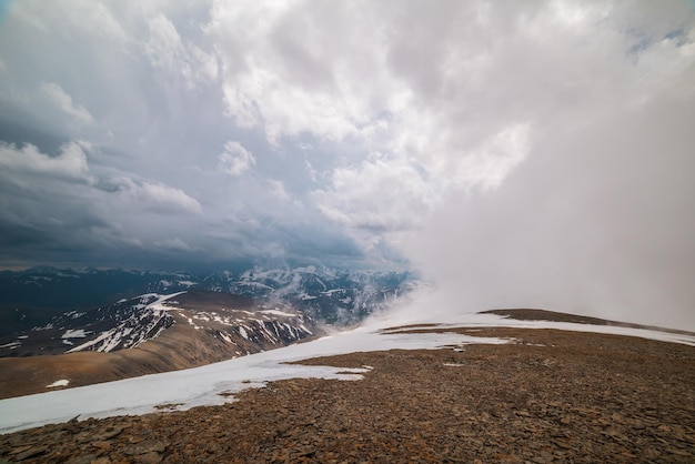 Awesome top view through clouds to high snowy mountains Scenic landscape with beautiful snow mountains in low clouds Atmospheric alpine view from stone hill to snow mountain range with low clouds