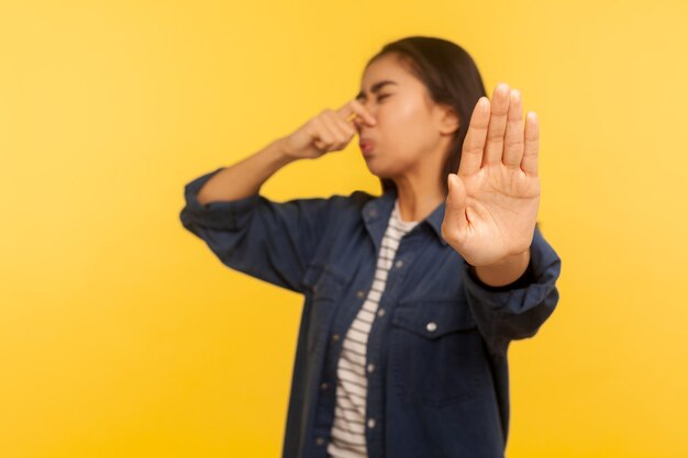 Foto odore terribile ritratto di ragazza dispiaciuta in camicia di jeans che fa una smorfia di disgusto e trattiene il respiro afferrando il naso e mostrando stop per evitare odore puzzolente scoreggia gas indoor studio girato sfondo giallo