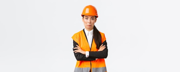 Awkward asian female engineer in reflective jacket safety helmet cross arms chest and smirk surprised making silly mistake acting natural standing white background
