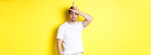 Awkward guy showing loser sign on forehead looking sad and gloomy standing in white tshirt against y