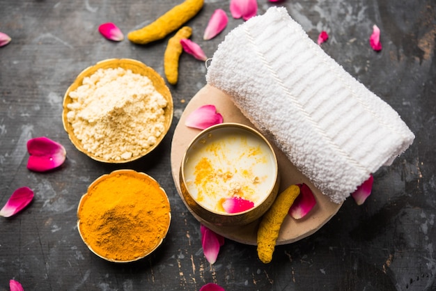 Ayurvedic face pack or mask using gram flouror besan, haldi & milk ubtan placed in brass bowls with rose petals, scrub and white napkin. selective focus