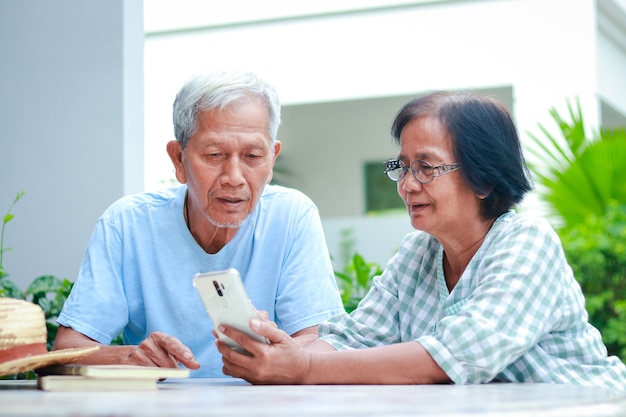 Aziatisch bejaarde echtpaar dat gelukkig thuis woont zittend in de tuin met een smartphone, online chattend met uw kind. familieconcept, gezondheidszorg voor ouderen in de pensioengerechtigde leeftijd