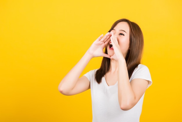 Aziatisch gelukkig portret mooie schattige jonge vrouw tiener staande grote schreeuw met handen naast mond geven opgewonden positief kijkend naar kant geïsoleerd, studio opname op gele achtergrond met kopie ruimte