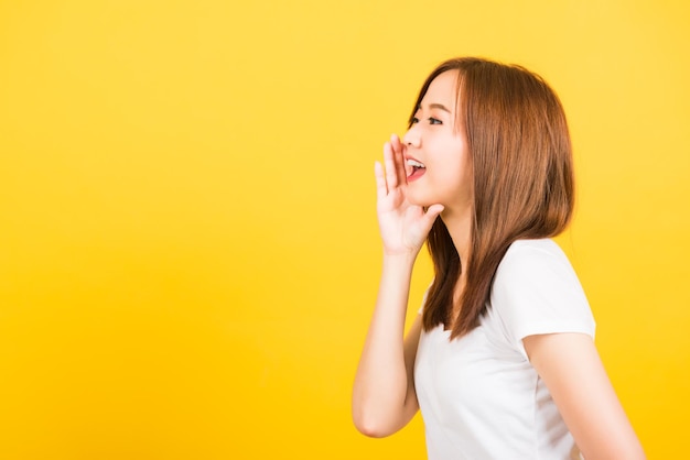 Aziatisch gelukkig portret mooie schattige jonge vrouw tiener staande slijtage t-shirt hand op mond praten fluisterend geheim gerucht op zoek naar kant geïsoleerd, studio opname op gele achtergrond met kopie ruimte