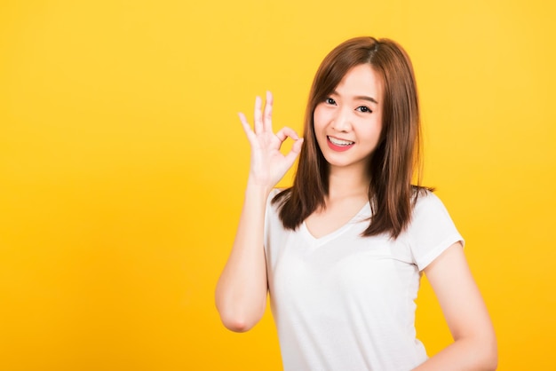 Aziatisch gelukkig portret mooie schattige jonge vrouw tiener staande slijtage t-shirt tonen gebaren ok teken met vingers kijken naar camera geïsoleerd, studio opname op gele achtergrond met kopie ruimte voor tekst