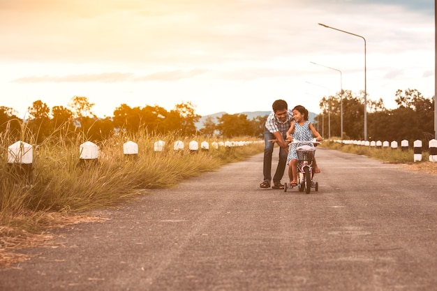 Aziatisch jong geitjemeisje die pret hebben om fiets met vaderhulp te berijden om hen samen te duwen