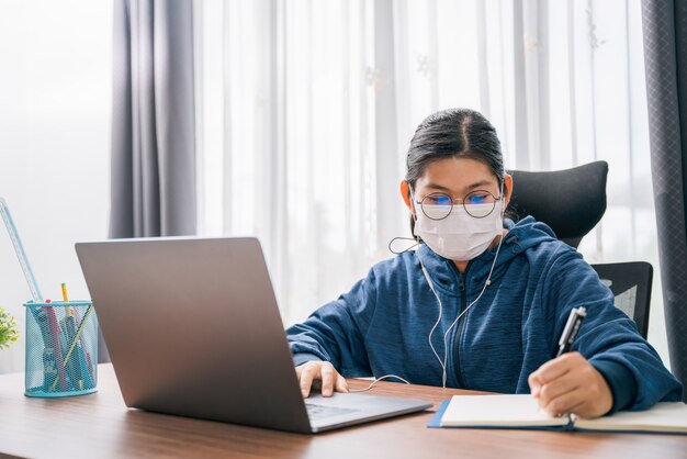 Aziatisch jong meisje draagt een masker student met een bril hoofdtelefoon meisje studie gelukkig schrijven notitie over een boek op zoek videoconferentie laptopcomputer online internet leren afstandsonderwijs thuis