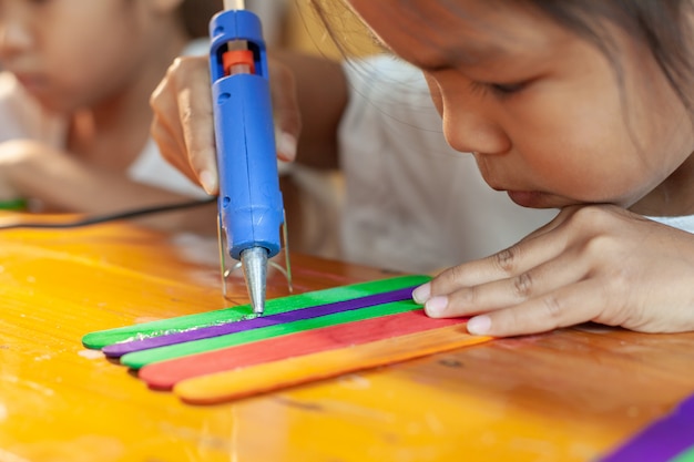 Aziatisch kindmeisje die gekleurde roomijsstokken lijmen door heet lijmpistool. kinderen hebben plezier om huis te maken in een handwerkproject.