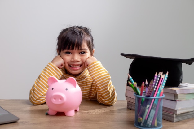 Aziatisch klein meisje met een afstudeerpet met een roze spaarvarken Geld besparen investeren in de toekomst