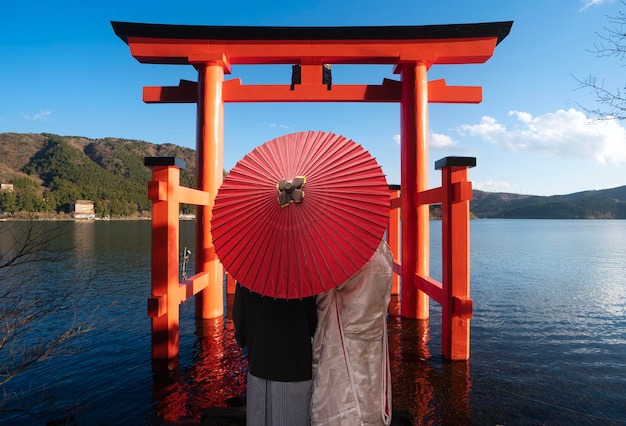 Aziatisch koppel in kimono trouwjurk staan samen met rode paraplu en rode torii poort