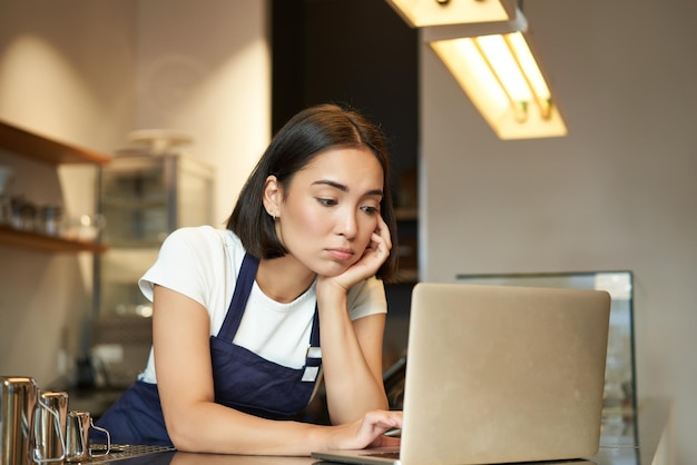 Aziatisch meisje barista staat achter de toonbank in café en kijkt naar een laptop die werkt terwijl ze zich depressief voelt