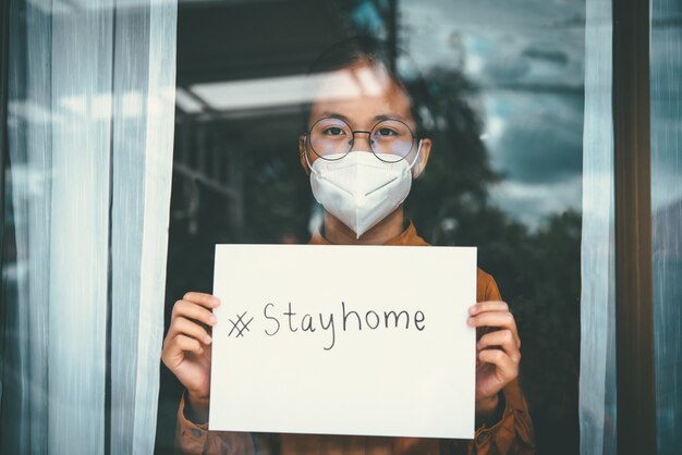 Aziatisch meisje dat een bril draagt, een masker draagt, papier met het label #stayhome vasthoudt, verveelt zich omdat ze de ziekte alleen thuis moet vasthouden en behandelen. Concept thuisquarantaine, preventie COVID-19