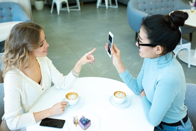 Aziatisch meisje foto's in smartphone tonen aan haar vriend wijzend op display tijdens discussie door kopje koffie in café