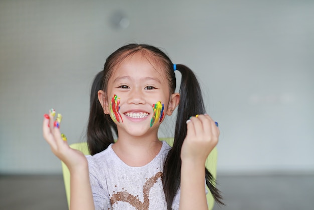 Aziatisch meisje met haar kleurrijke handen en wang geschilderd in de kinderkamer.
