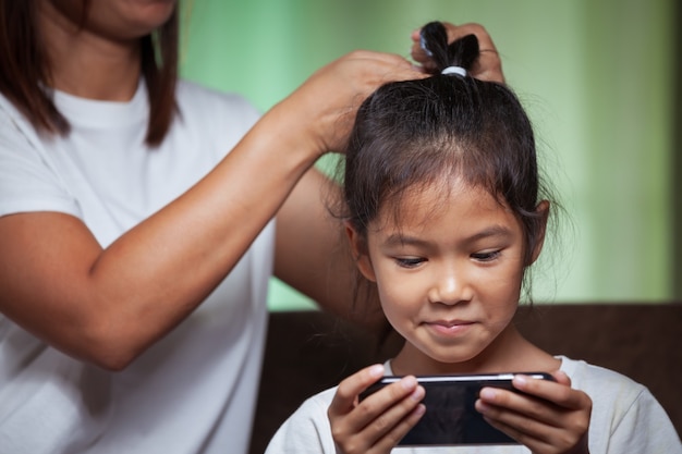 Aziatisch meisje spelen op telefoon terwijl moeder kapsel maakt