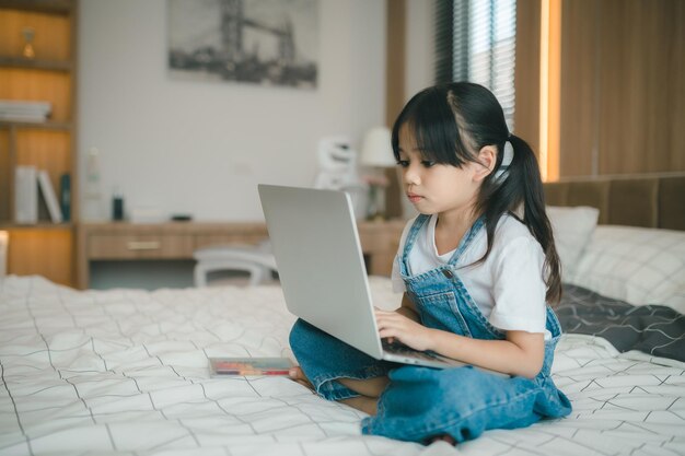 Aziatisch meisjeskind dat in bed zit en online leert op laptop Internet Virtuele les op video tijdens zelfisolatie thuis Video-onderwijs op afstand op afstand Moderne schoolstudie voor kinderen