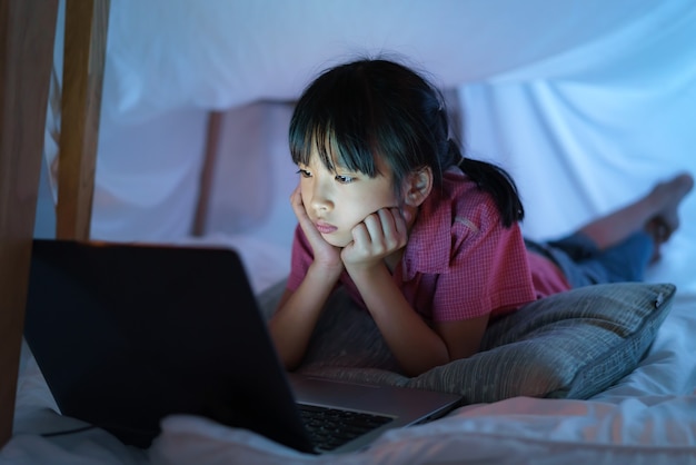 Aziatisch meisjeskind om een kamp te maken om fantasierijk te spelen, kijkend naar een film op laptop in de duisternis van het kamp in de huiskamer.