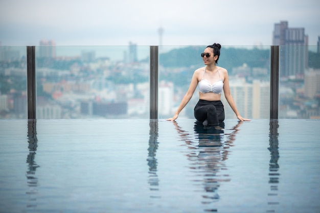 Aziatisch reisconcept. jonge vrouw die geniet van het uitzicht op de stad vanuit het zwembad op het dak van het hotel, de levensstijl van een mooi meisje buiten in vakantietijd