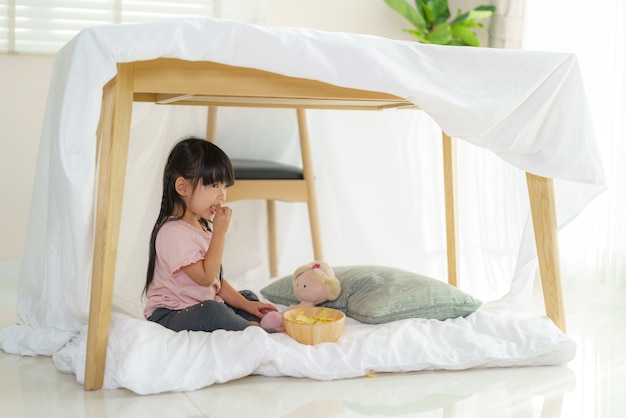 Aziatisch schattig klein meisje eten snack zittend in een deken fort in de huiskamer