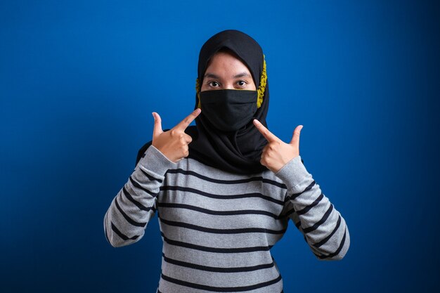 Aziatisch studentmeisje duim omhoog en maskers dragen om de verspreiding van het Corona-virus tegen een blauwe achtergrond te voorkomen terwijl ze terug naar school gaat.