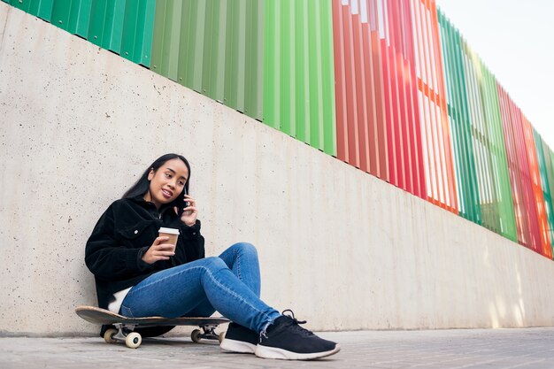 Aziatisch tienermeisje dat buiten aan de telefoon praat terwijl ze op haar skateboard tegen een betonnen muur leunt