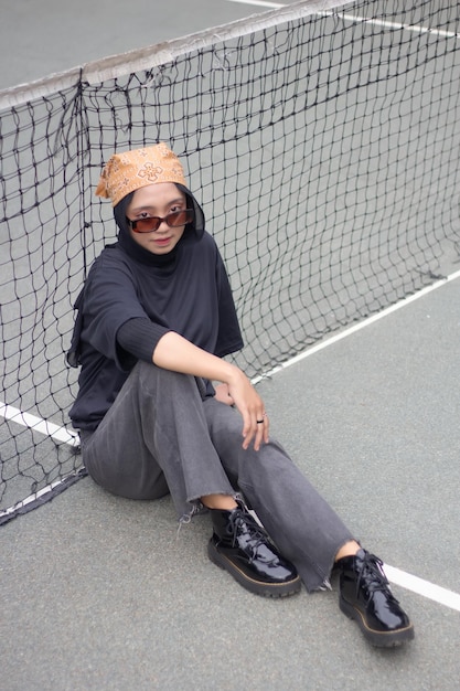 Aziatisch vrouwelijk model in jeans en een zwart shirt zit op een tennisbaan met een bandana en een bril