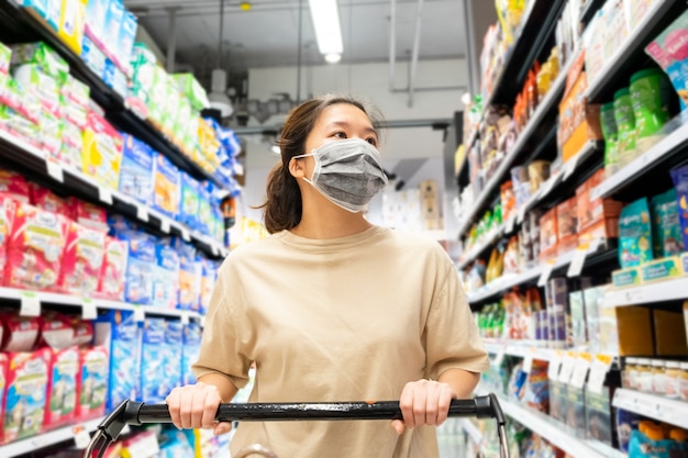 Aziatisch wijfje dat beschermend gezichtsmasker met boodschappenwagentje draagt bij supermarkt