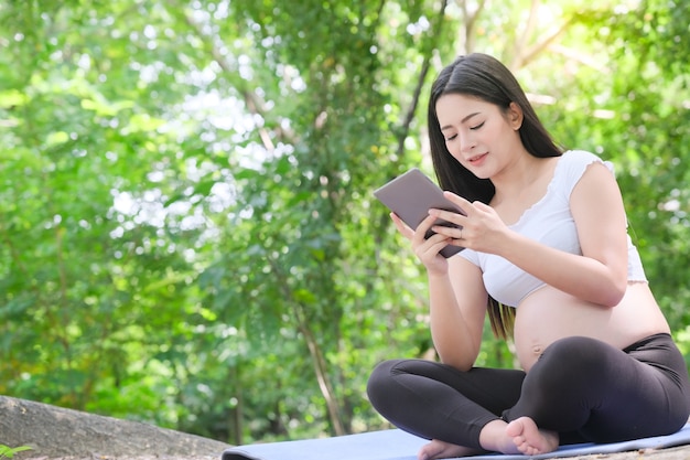 Aziatische 8 maanden zwangere vrouw die digitale tablet gebruikt die op gras in openluchtpark zit. Buiten natuur achtergrond.