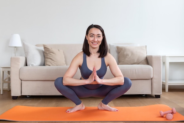 Aziatische aantrekkelijke lachende vrouw beoefenen van yoga zitten in Half Lotus oefening Ardha Padmasana pose uitwerkend dragen van sportkleding meditatie sessie binnen volledige lengte interieur