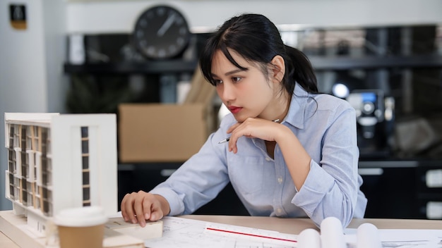 Aziatische architecten en ingenieursvrouwen die werken aan het onderzoeken van het ontwerp van architectonische bouwmodellen en het maken van aantekeningen op papier tijdens het plannen van de bouw en het architectonische projectplan op blauwdruk in het kantoor