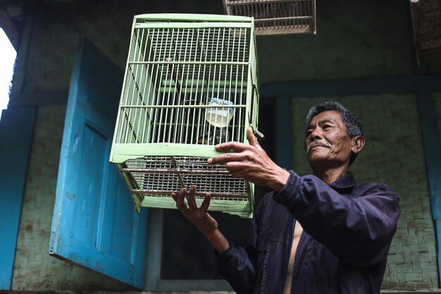 Aziatische bejaarde die groene vogelkooi buiten traditioneel houten huis houdt