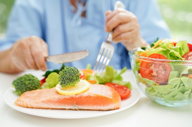 Aziatische bejaarde patiënt die zalmstaak en groentesalade eet voor gezond voedsel in het ziekenhuis