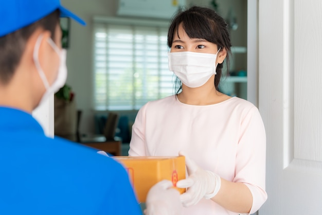 Aziatische bezorger die gezichtsmasker en handschoen in blauw uniform draagt met kartonnen dozen vooraan in huis en vrouw die een levering van dozen van bezorger accepteert tijdens COVID-19-uitbraak.