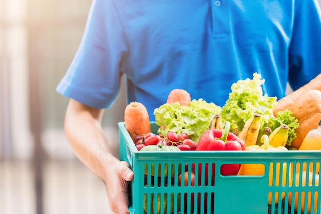 Aziatische bezorger van de kruidenierswinkel in blauw uniform en gezichtsmasker beschermt hij die verse groenten in plastic doos aan de deur thuis bezorgt na de uitbraak van het coronavirus, terug naar het nieuwe normale concept